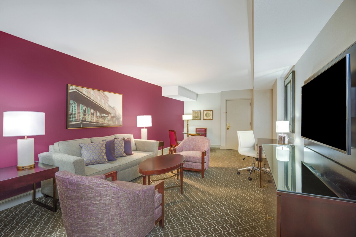 Ein Hotelzimmer mit roter Akzentwand, einem großen Flachbildfernseher, einem Sofa, zwei Sesseln, einem Couchtisch und einem Schreibtisch mit Lampe.