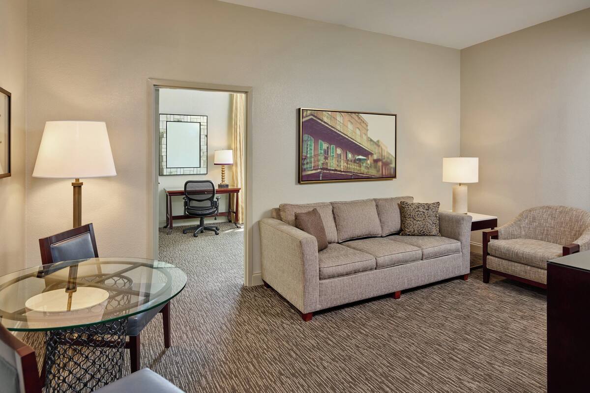 Quarto de hotel com mesa de jantar de vidro, sofá bege, duas poltronas, um quadro na parede e chão coberto por carpete.