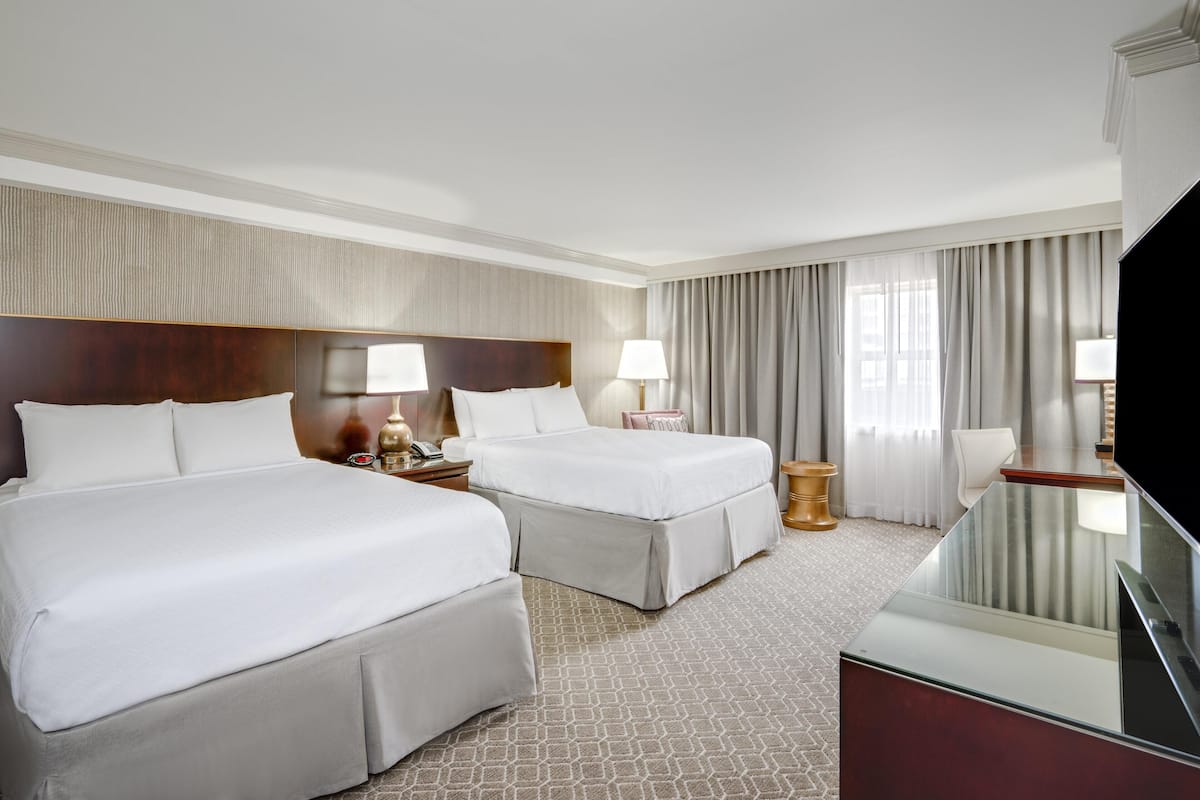 A hotel room with two beds, a TV, a desk, and a large window with curtains.