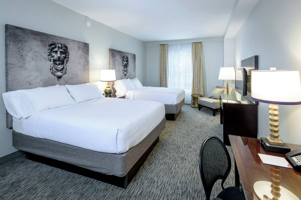 Quarto de hotel com duas camas, uma escrivaninha, uma cadeira, televisão e obras de arte nas paredes.