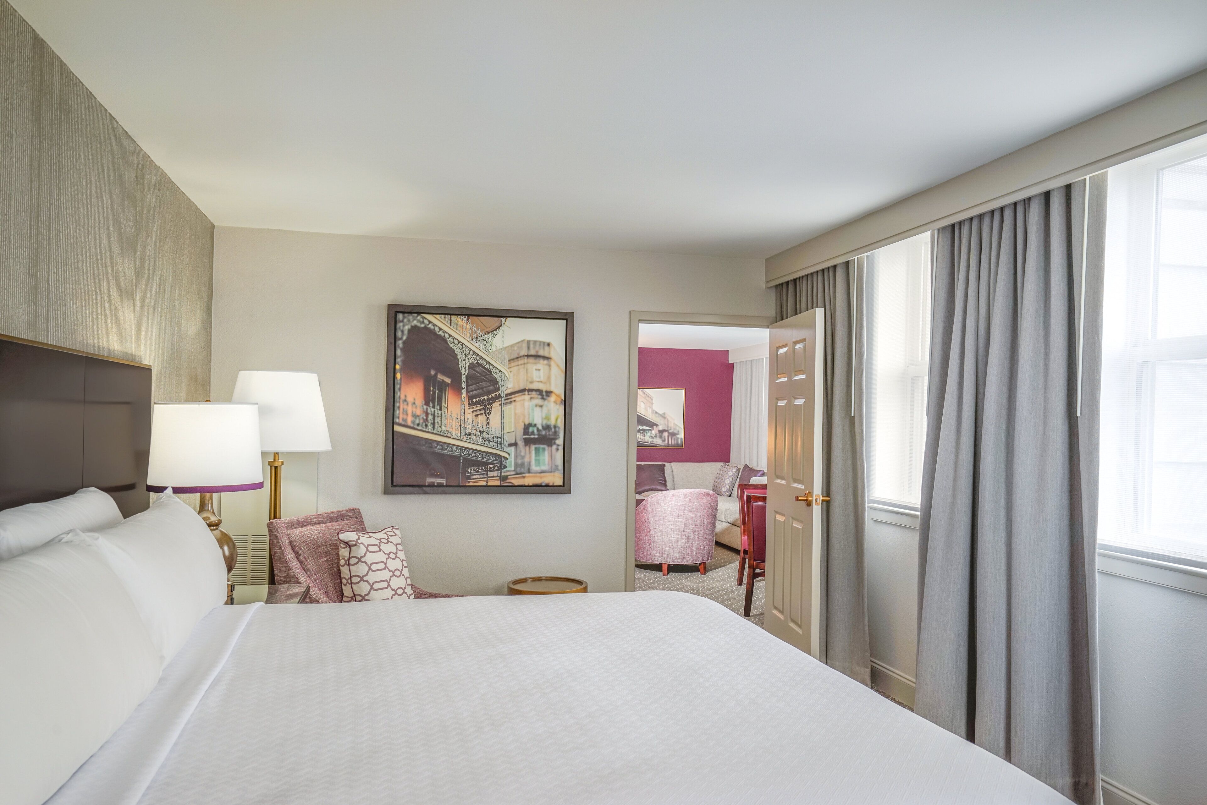 A hotel room with a large bed, two bedside lamps, a framed cityscape painting, a small round table, and a view of the outside through a window with curtains.