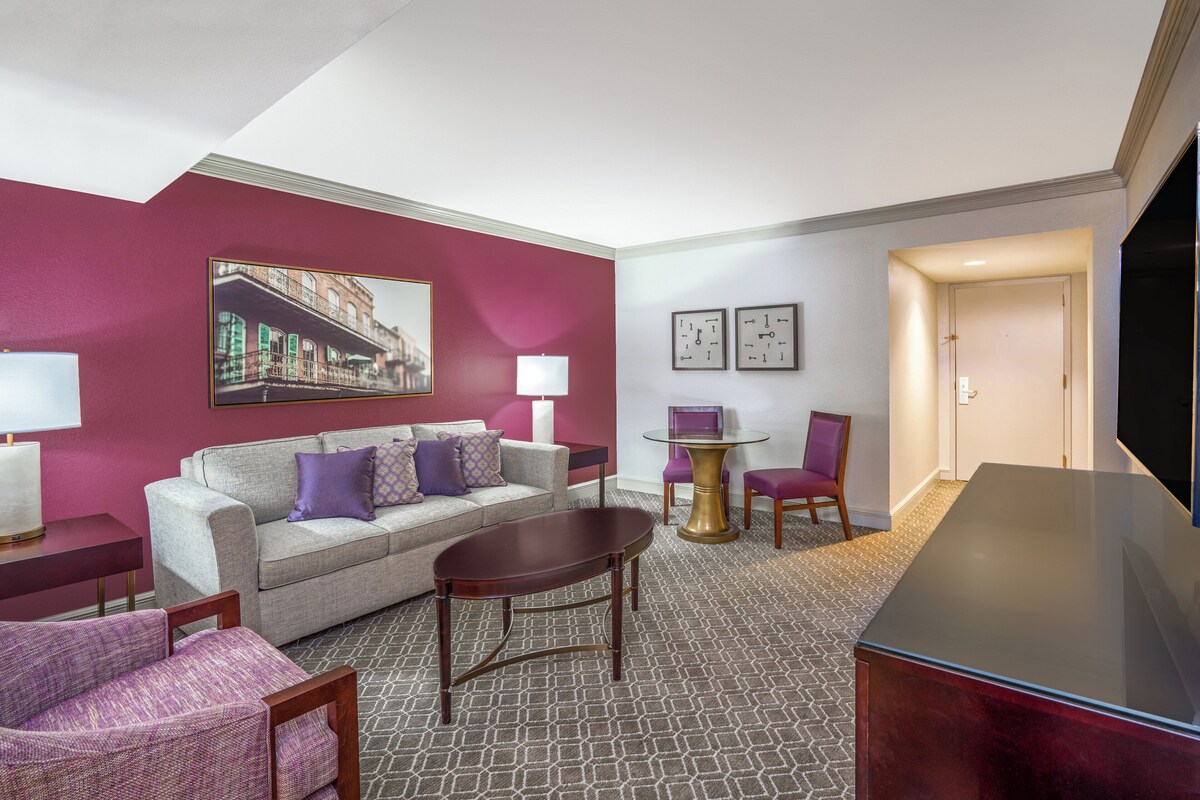 Ein Hotelzimmer mit grauem Sofa, einem violetten Sessel, einem Holztisch, einem Fernseher und einem Bild an der Wand.