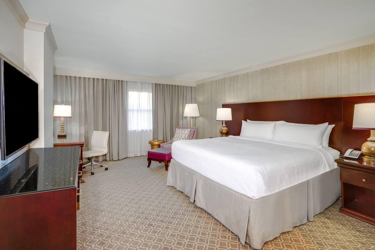 Quarto de hotel com uma cama grande, uma escrivaninha com televisão de tela plana, uma cadeira, duas luminárias de cabeceira e uma janela ampla com cortinas.