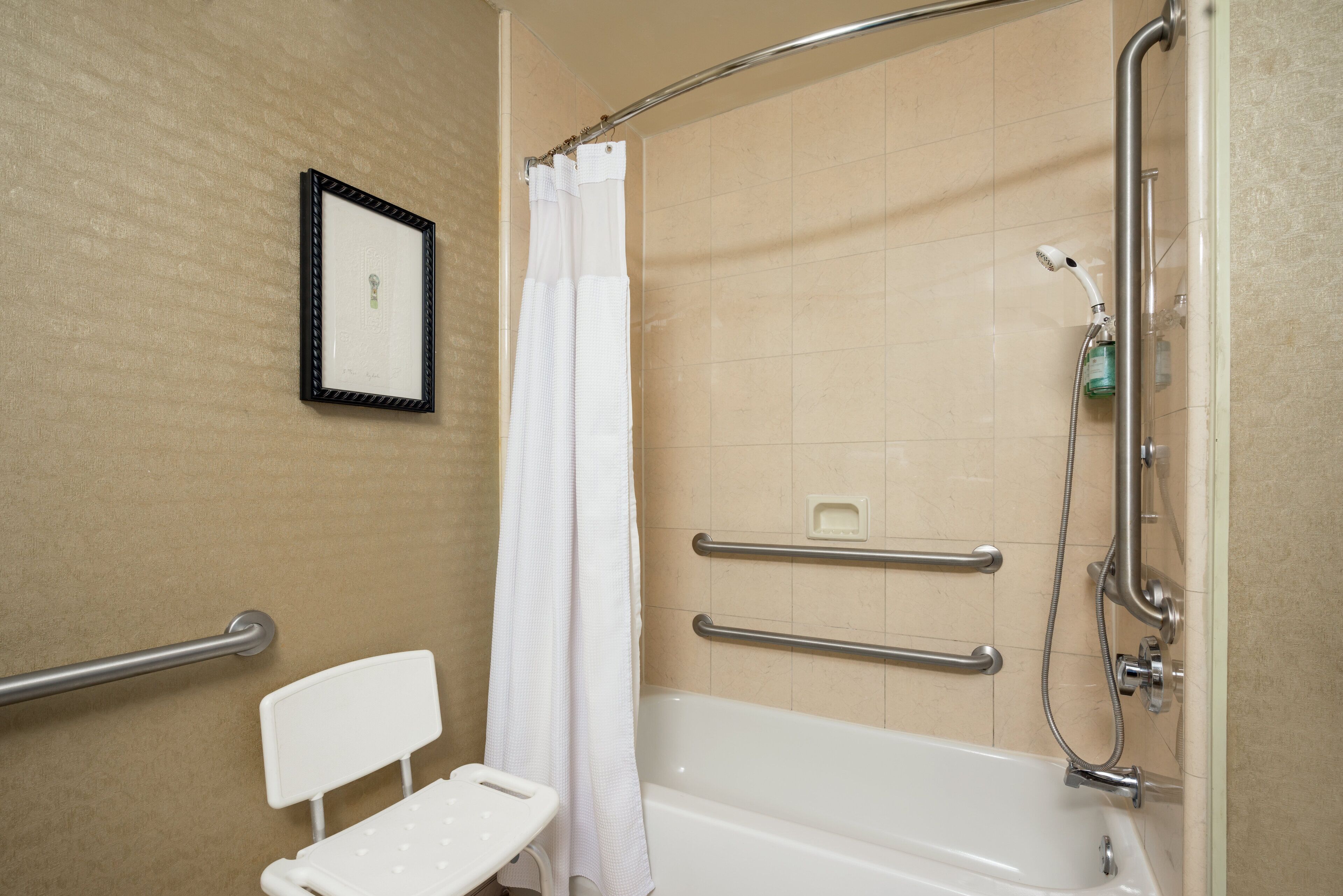 Baño con ducha, barras de apoyo, un banco blanco y un cuadro en la pared.