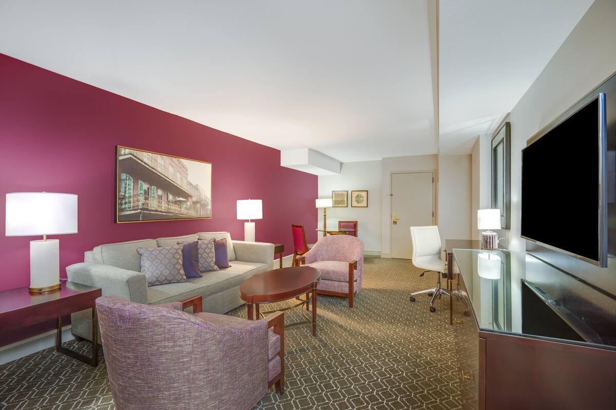 Quarto de hotel com parede em vermelho, uma televisão de tela plana grande, um sofá, duas poltronas, uma mesa de centro e uma escrivaninha com luminária.