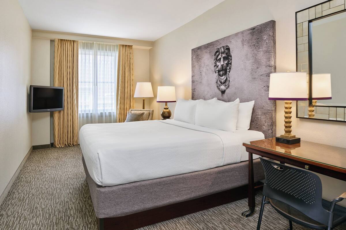 Quarto de hotel com uma cama grande, duas luminárias de cabeceira, uma TV de tela plana, uma escrivaninha com cadeira e um quadro abstrato de grandes dimensões na parede.