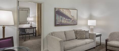 A hotel room with a beige sofa, a glass table, a chair, a desk with a lamp, and a framed picture on the wall.