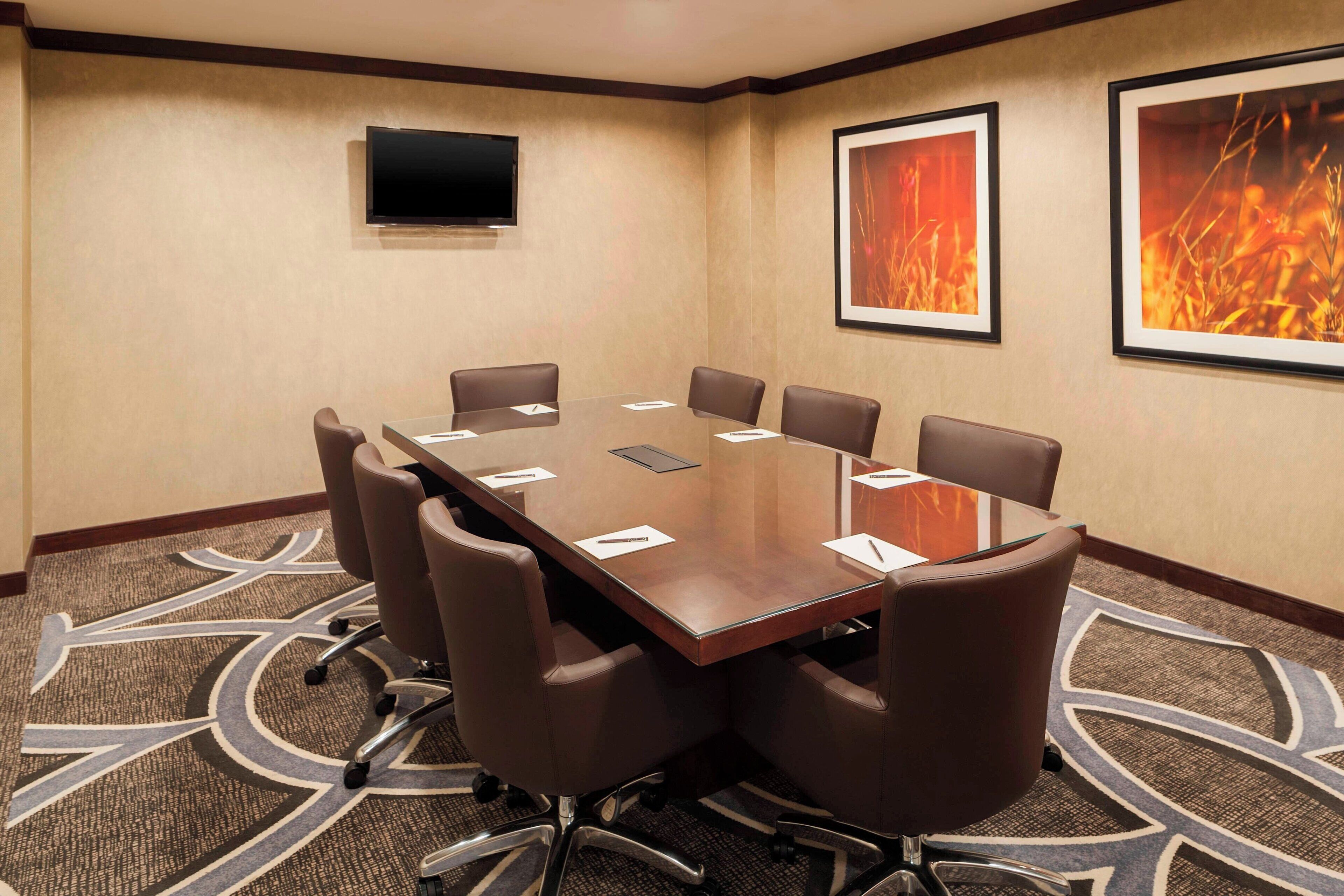 A conference room with a large table, eight chairs, a flat-screen TV, and framed artwork on the walls.