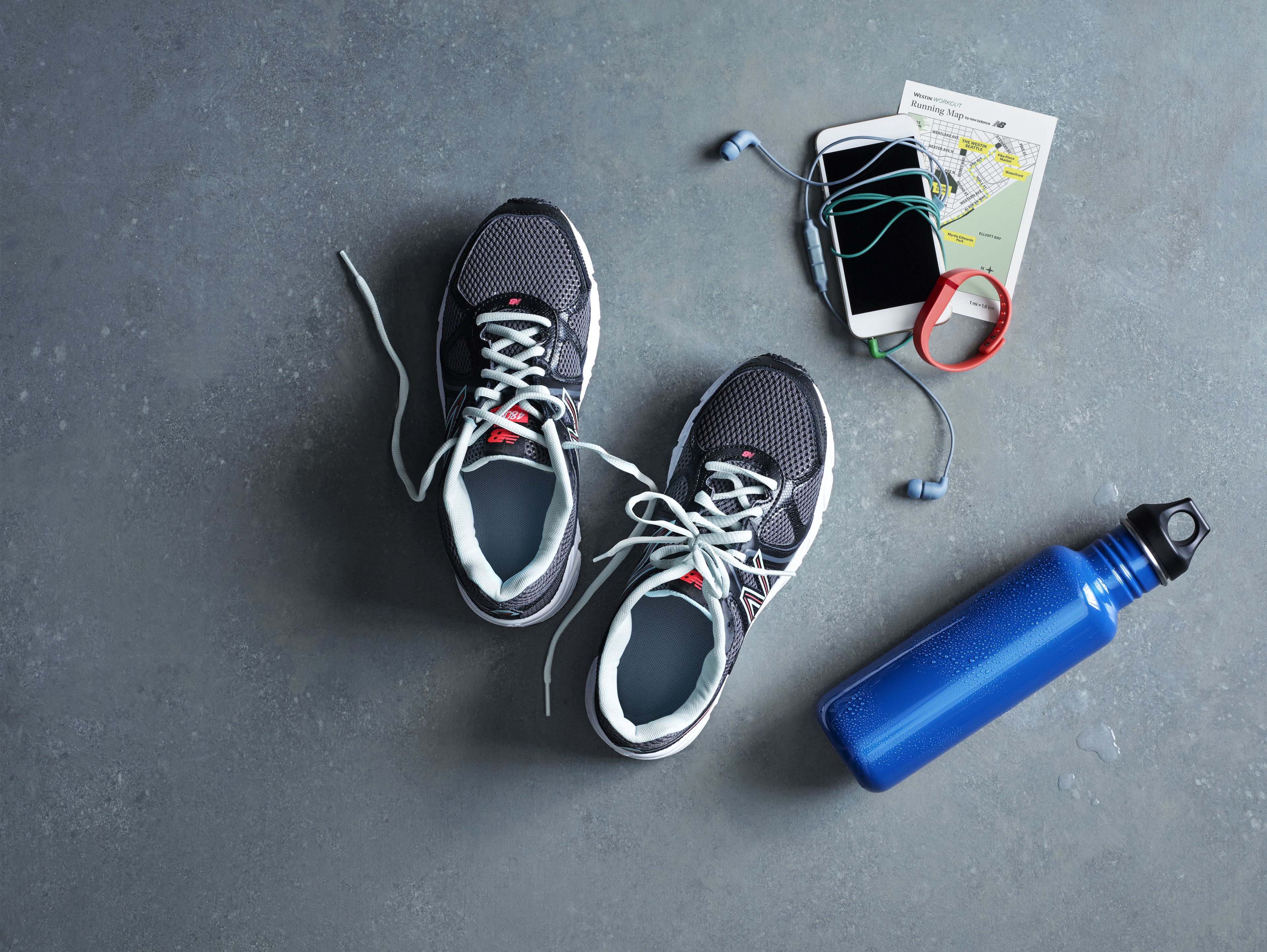 A pair of sneakers, a smartphone, earphones, a map, and a water bottle on a grey surface.