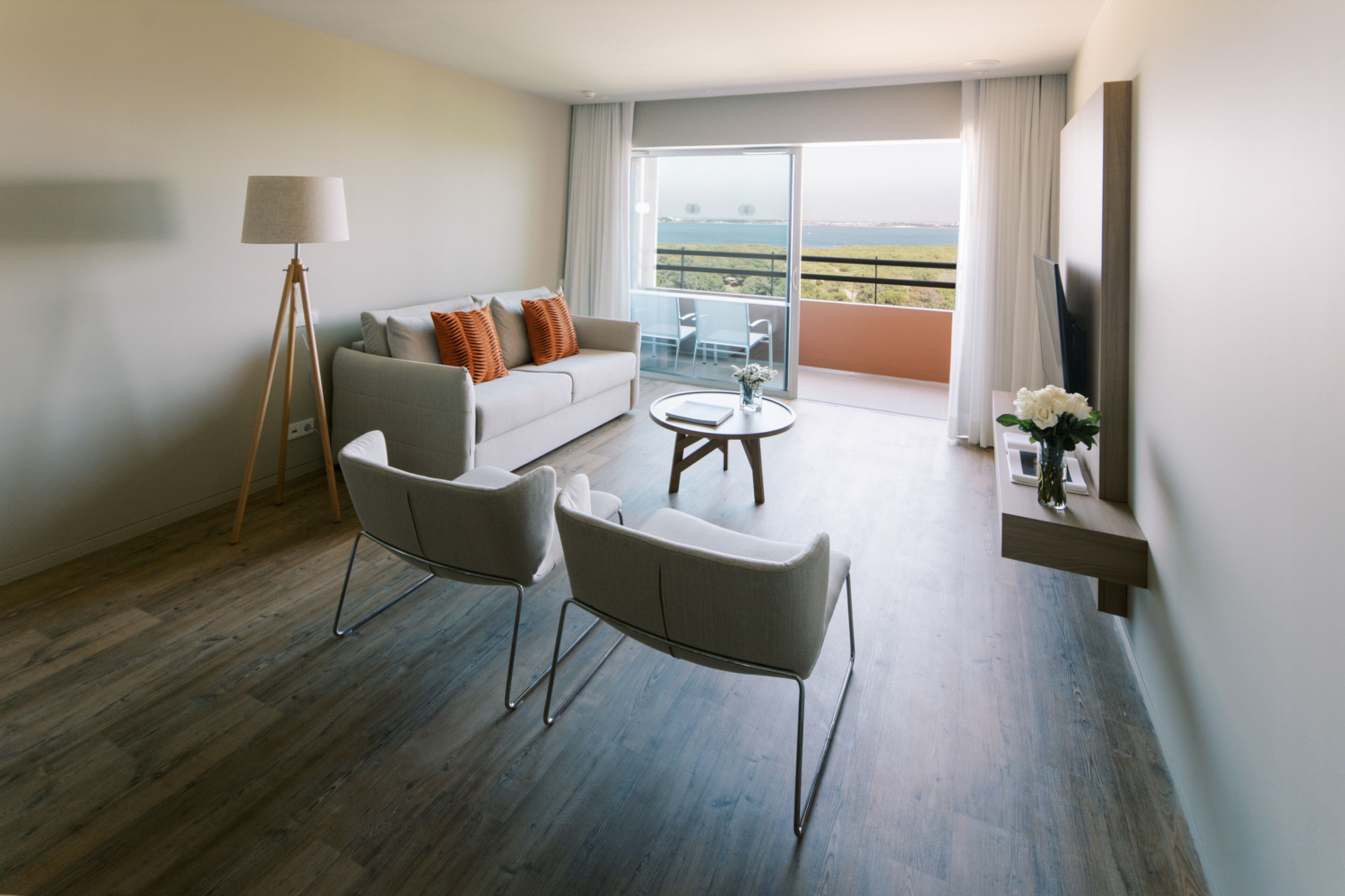 A modern hotel room with a sofa, two armchairs, a small table, a floor lamp, a TV, and a balcony with a view.