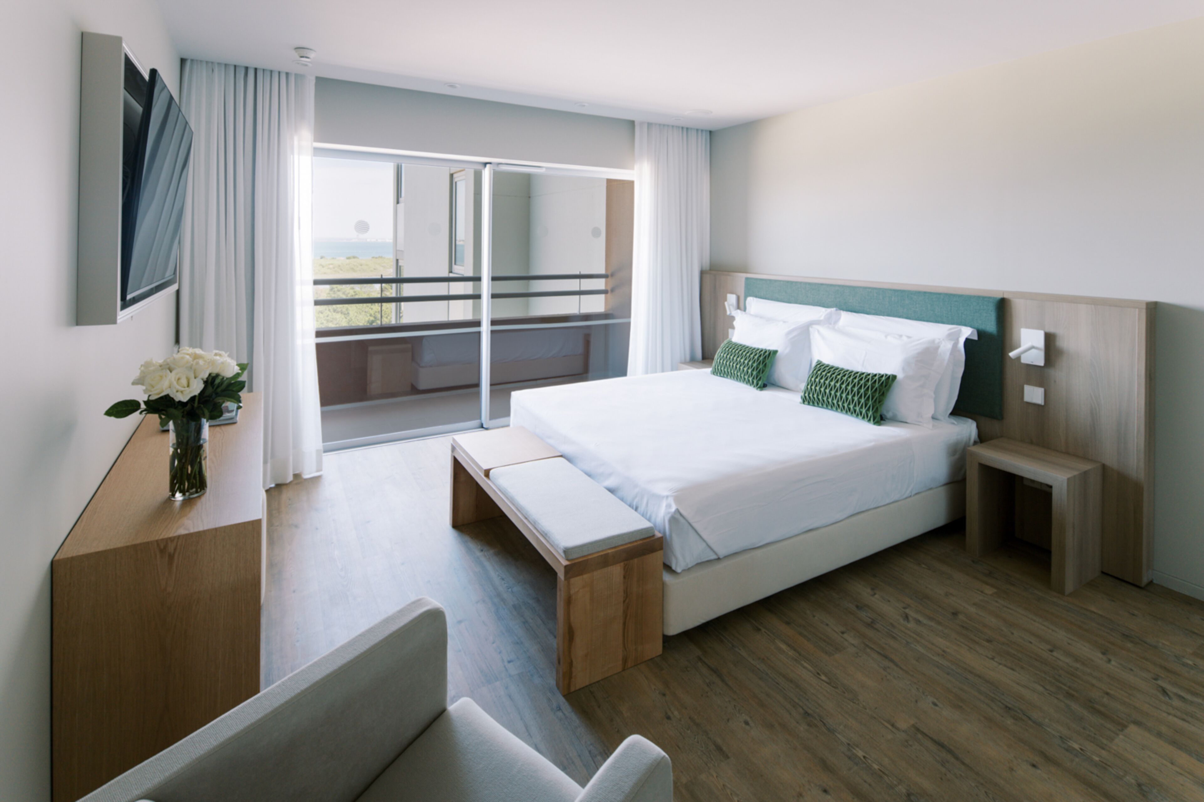 A modern hotel room with a large bed, a wooden bench, a bedside table, and a view of the outdoors.