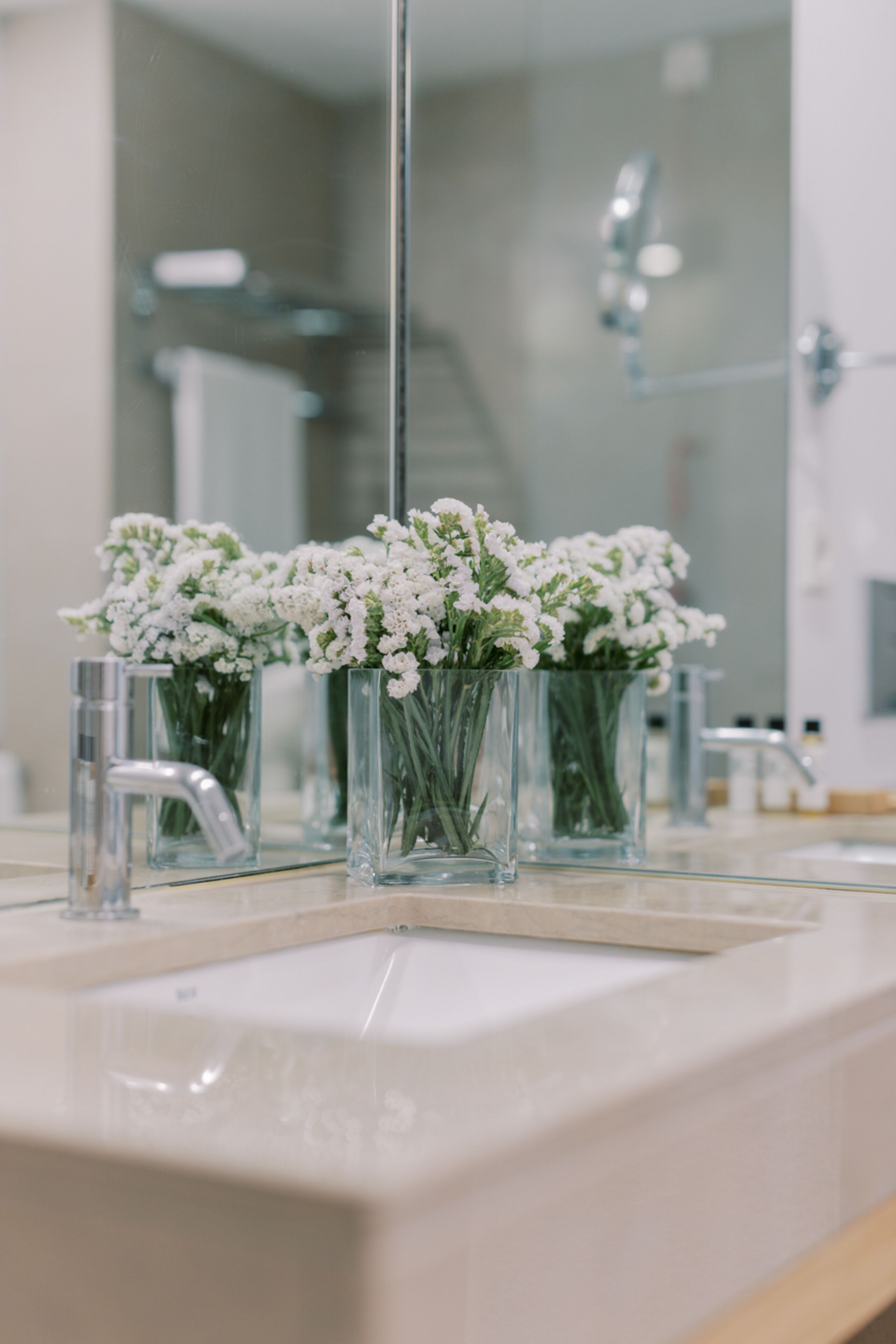 Casa de banho com lavatório branco, dois vasos de vidro com flores brancas e um espelho a refletir o espaço.