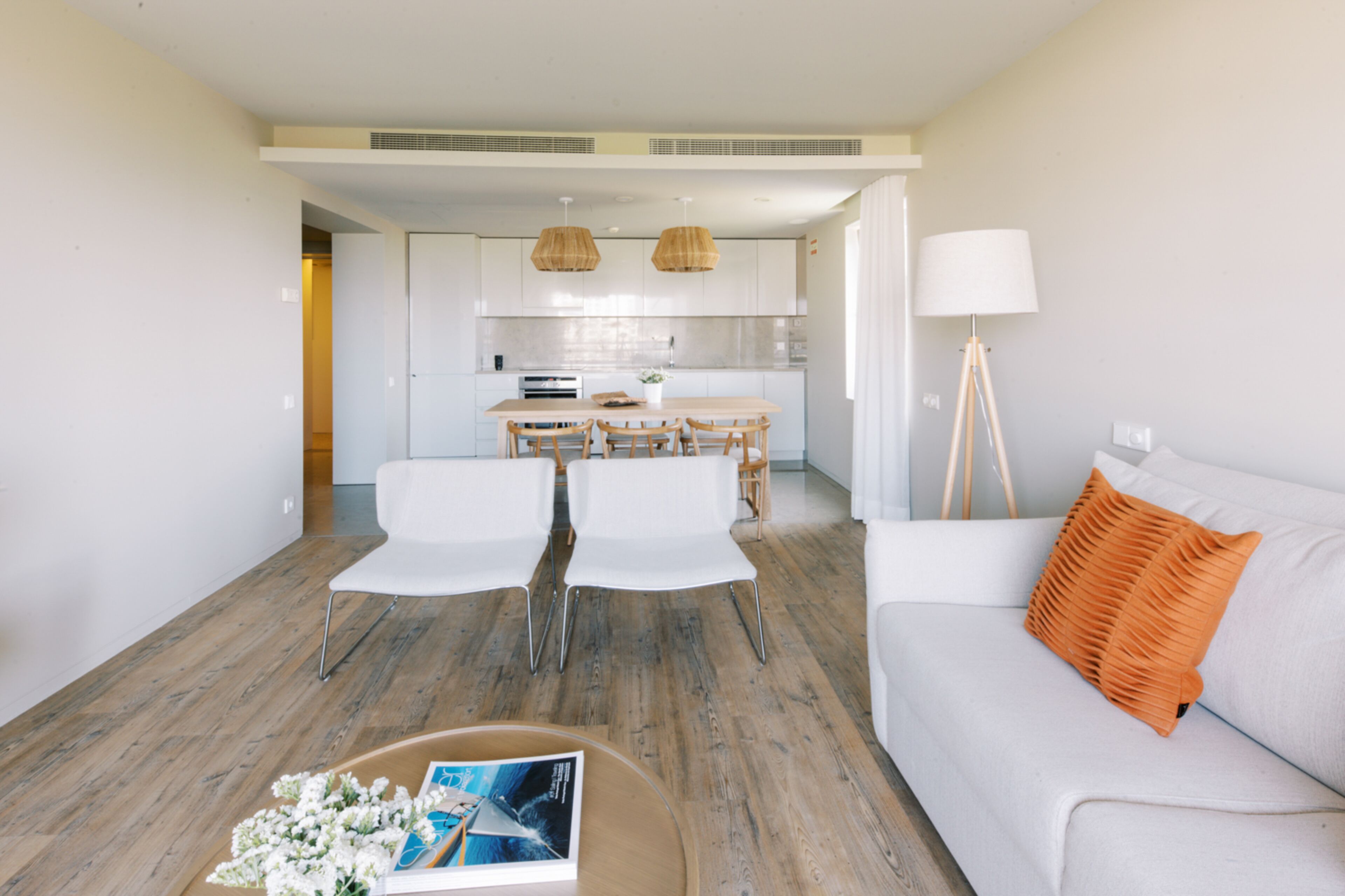 A modern living room with a sofa, a coffee table with books and flowers, and a dining area with chairs and a table.
