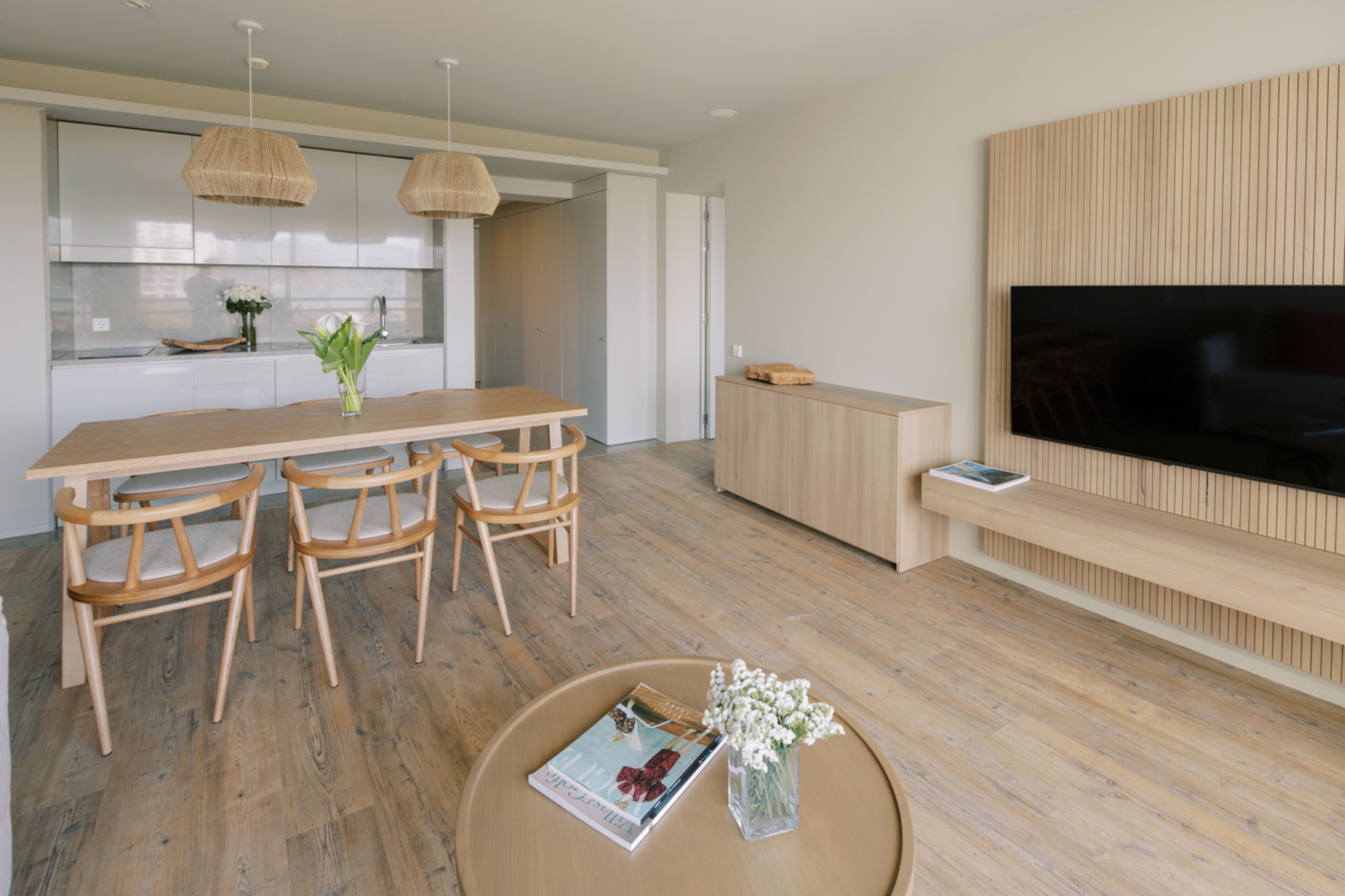 Een moderne keuken met witte kasten, een houten eettafel met stoelen en een grote flatscreen televisie aan de muur.