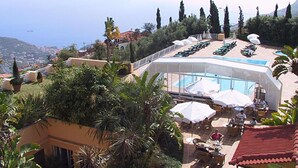 Un área de piscina en la azotea con sillones de descanso y sombrillas, rodeada de vegetación y con vistas a un pueblo costero.