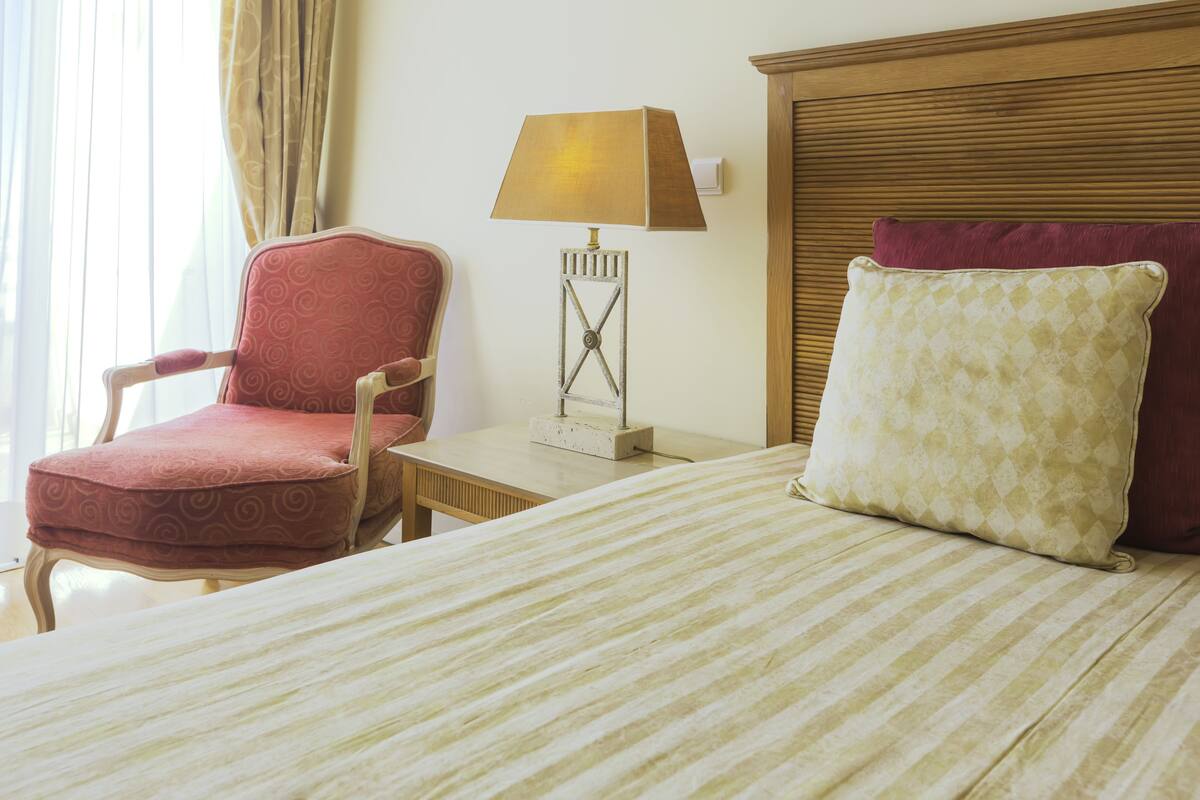 A hotel room with a bed, a red armchair, a small table, and a lamp.