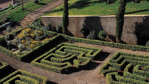 Un jardín ajardinado con patrones geométricos de setos, un edificio blanco con tejado rojo y vistas al mar al fondo.
