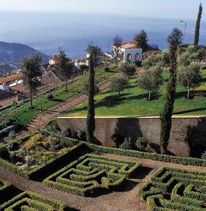 Un jardin aménagé avec des motifs géométriques créés à l’aide de haies, un bâtiment blanc avec un toit rouge et, au loin, une vue sur la mer.