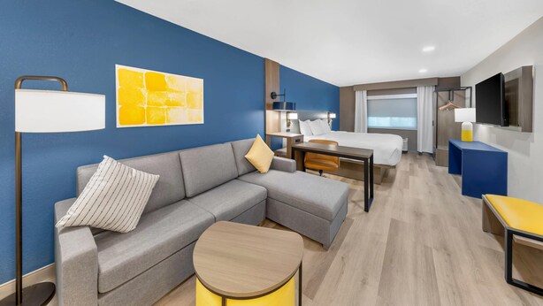 A modern hotel room with a grey sectional sofa, a small round table, a bed with a wooden headboard, a TV, and a large window. - Comfort Inn Fontana (Fontana)