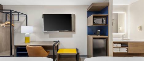 A hotel room with a bed, a desk with a chair, a television, a microwave, a mini-fridge, and a bathroom with a sink and mirror.