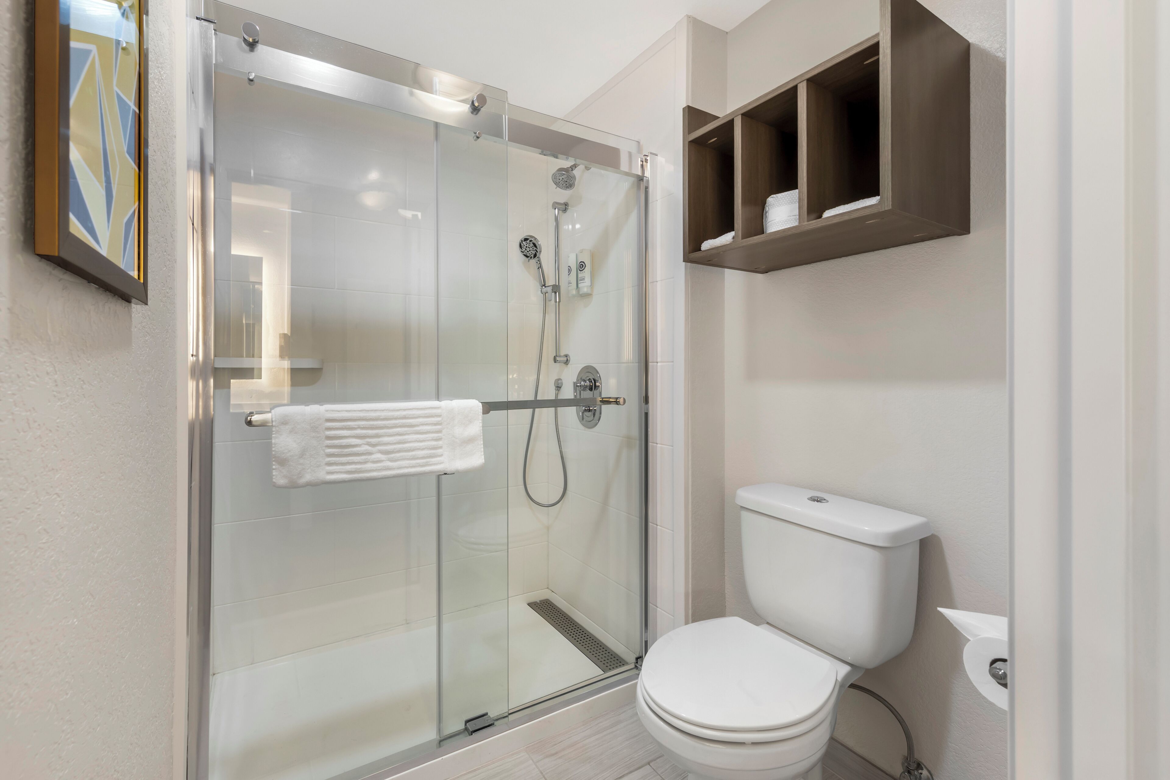 Un baño moderno con ducha envuelta en vidrio, inodoro blanco y un pequeño estante sobre él.