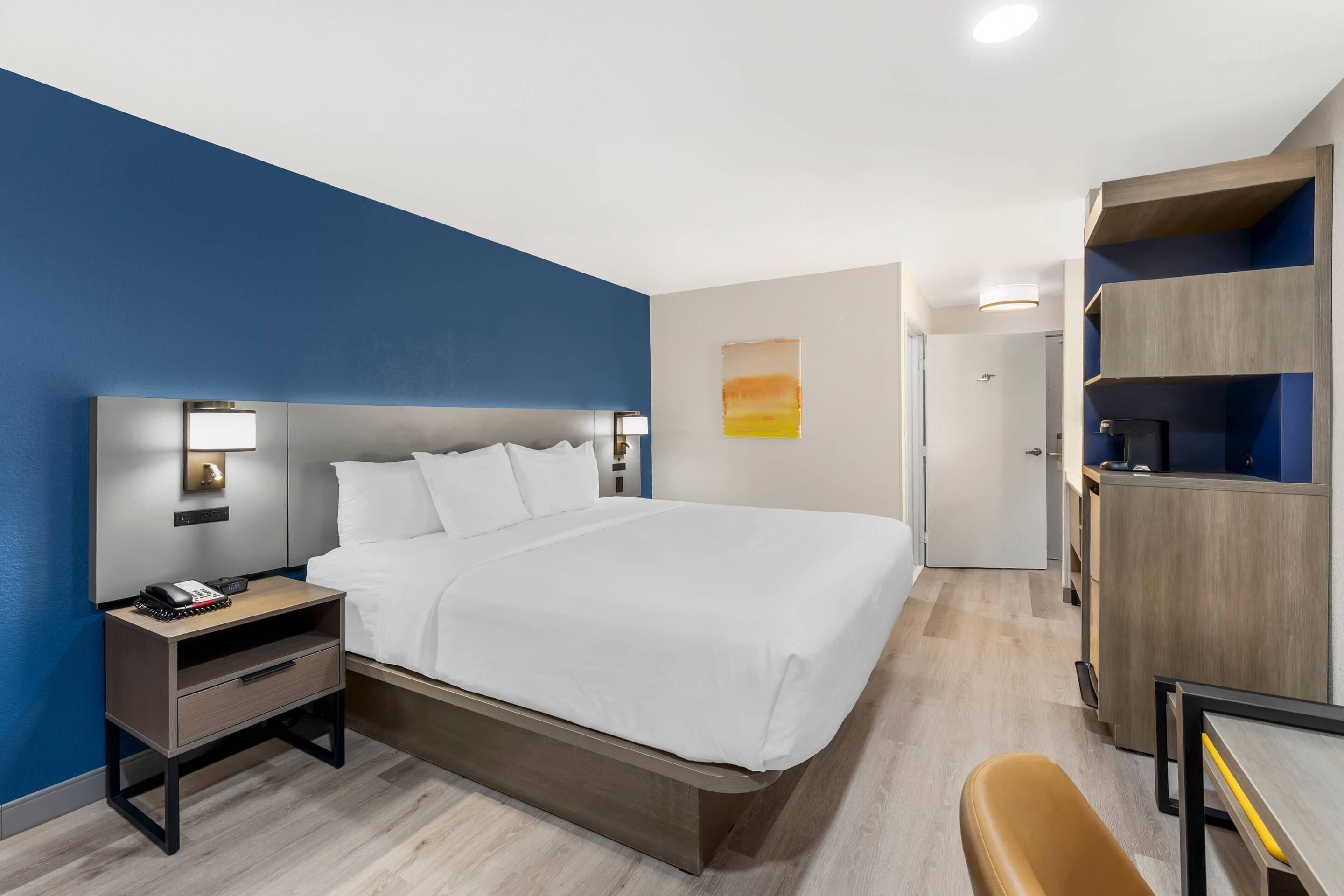 A hotel room with a bed, bedside table, lamp, and a desk with a chair.