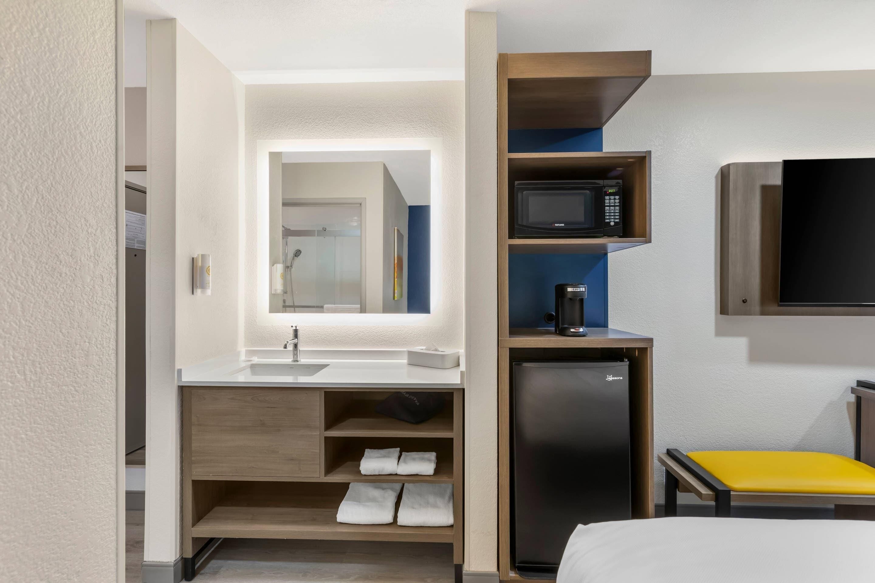 A hotel room with a bed, a small desk with a chair, a microwave, a refrigerator, and a TV.