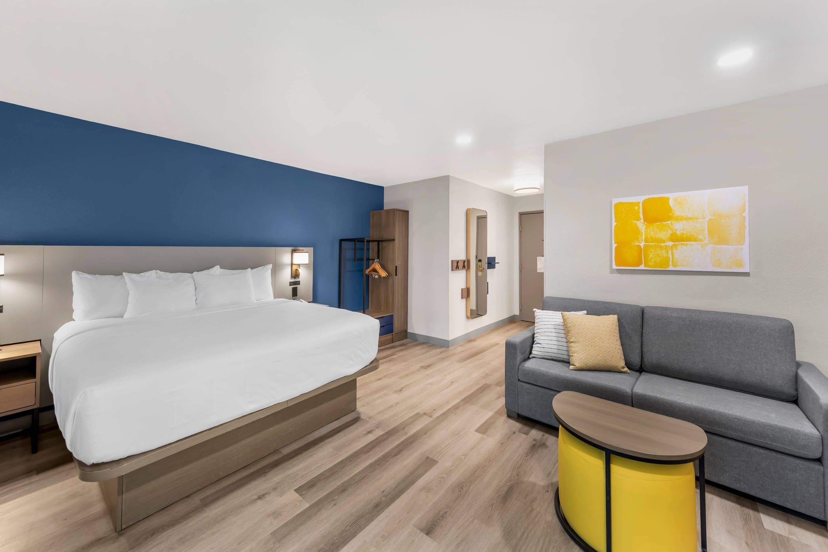 Una habitación de hotel con una cama grande, un sofá gris, una mesita auxiliar amarilla pequeña, un espejo y una obra de arte en la pared.