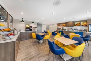 A modern dining area with yellow and blue chairs, wooden tables, and a kitchen in the background. - Comfort Inn Fontana (Fontana)