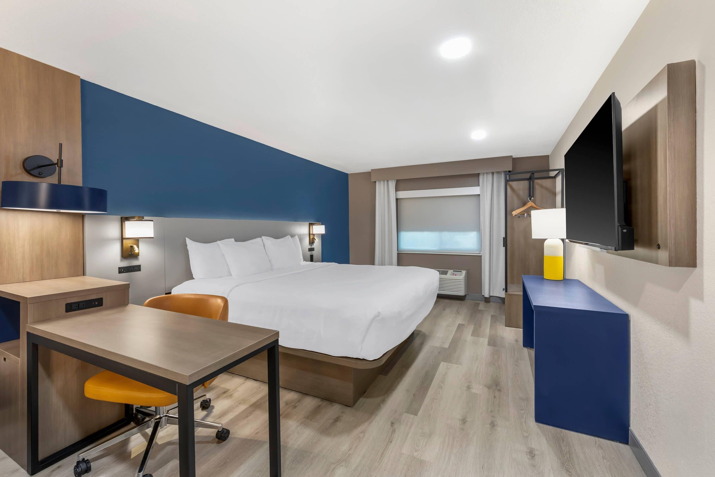 Una habitación de hotel con cama, escritorio, silla, televisión y ventana con cortinas.