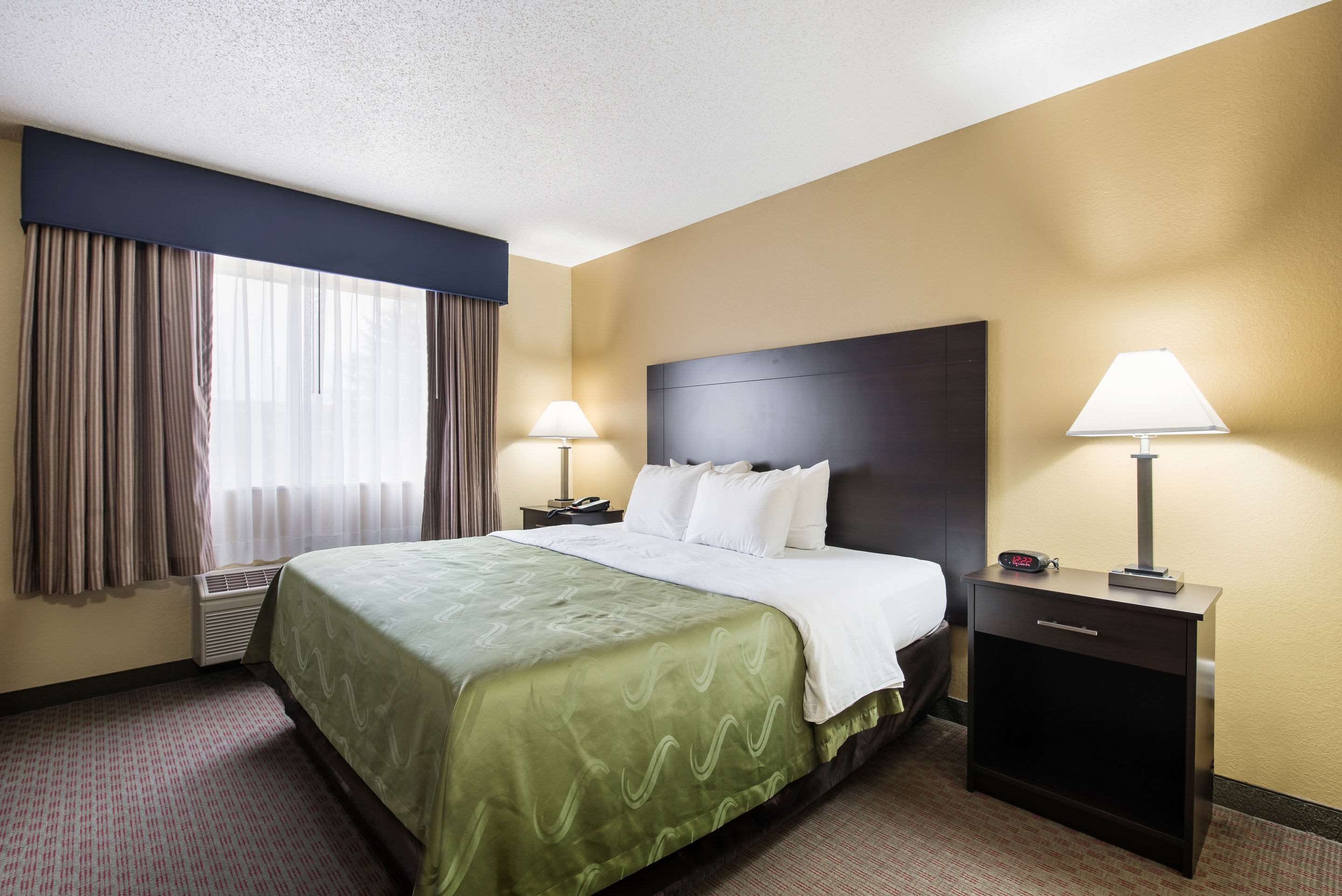 A hotel room with a large bed, two bedside tables with lamps, a headboard, and a window with curtains.
