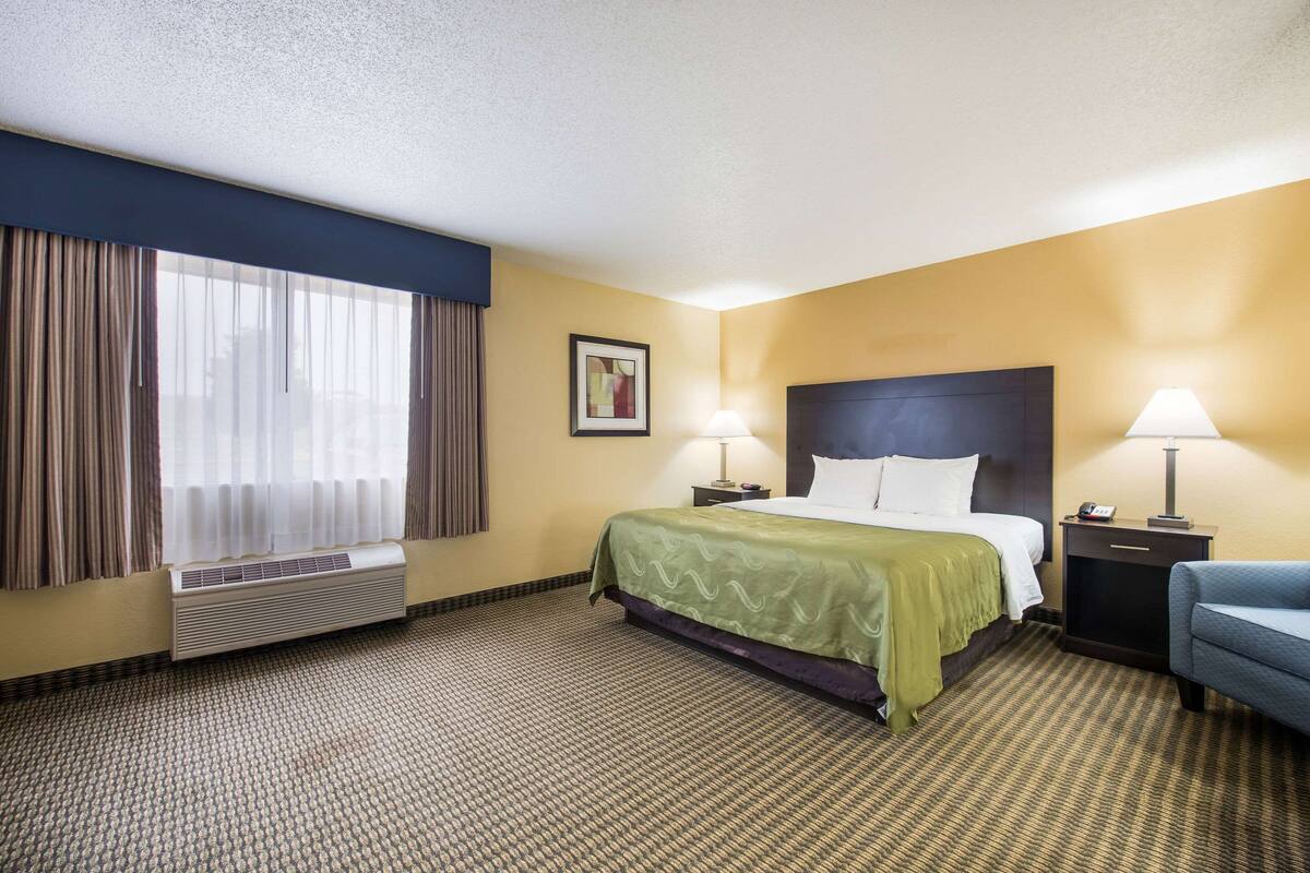 Quarto de hotel com cama, duas mesas de cabeceira com luminárias, uma janela com cortinas e ar condicionado fixado na parede.