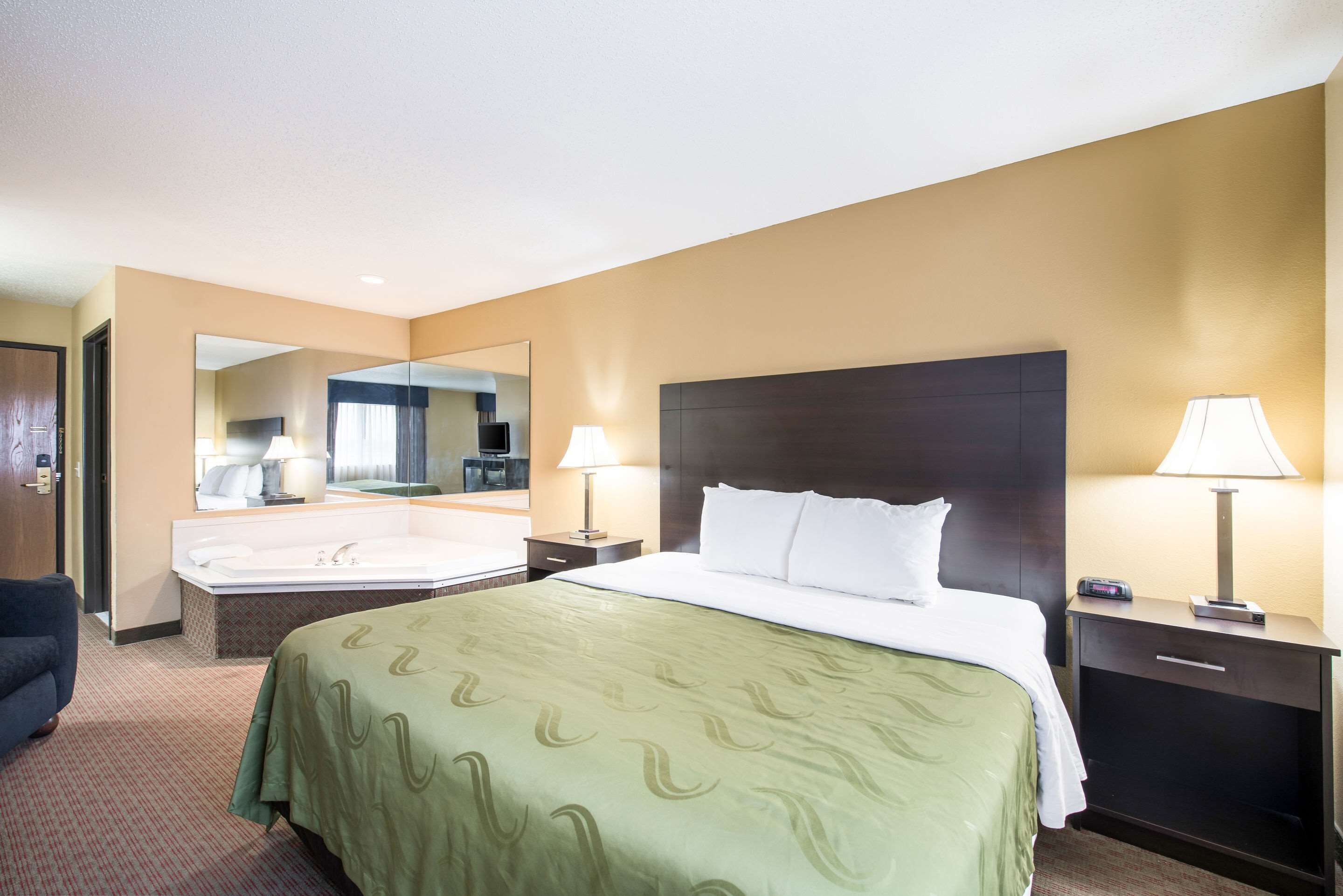 A hotel room with a large bed, a bedside table with a lamp, a mirror, and a view of the bathroom.