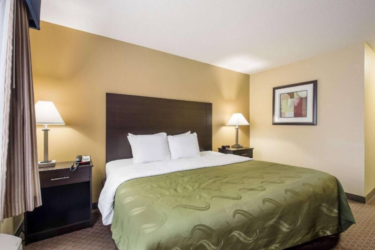 Ein Hotelzimmer mit einem Bett, zwei Nachttischen, einer Lampe auf jedem Tisch, einem gerahmten Bild an der Wand und einem halb heruntergezogenen Vorhang.