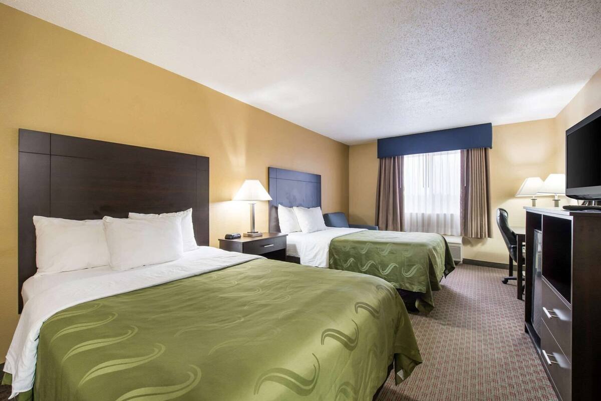 Quarto de hotel com duas camas, uma escrivaninha, televisão e janela com cortinas.