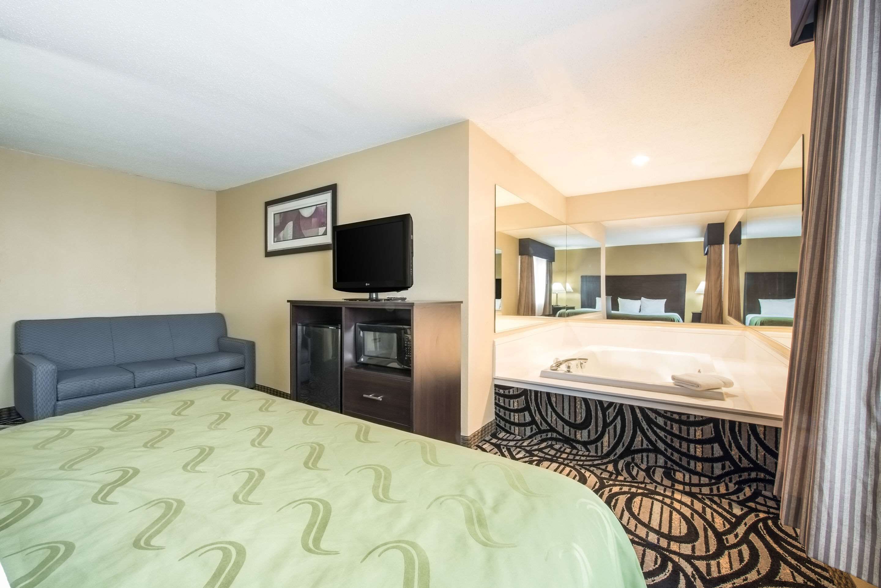 A hotel room with a bed, a television, a sofa, a desk with a microwave, and a bathroom with a sink and mirror.