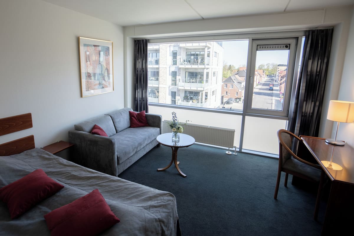 A hotel room with two beds, a sofa, a small table with a vase, and a view of buildings through large windows.
