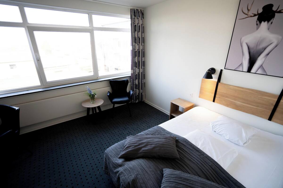 A hotel room with a large bed, a small table with a plant, a chair, a nightstand, and a window with curtains.