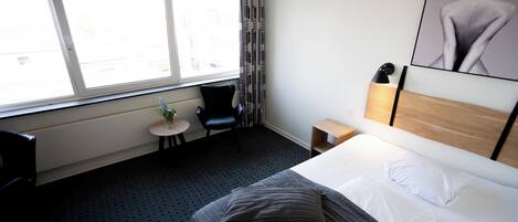 A hotel room with a large bed, a small table with a plant, a chair, a nightstand, and a window with curtains.