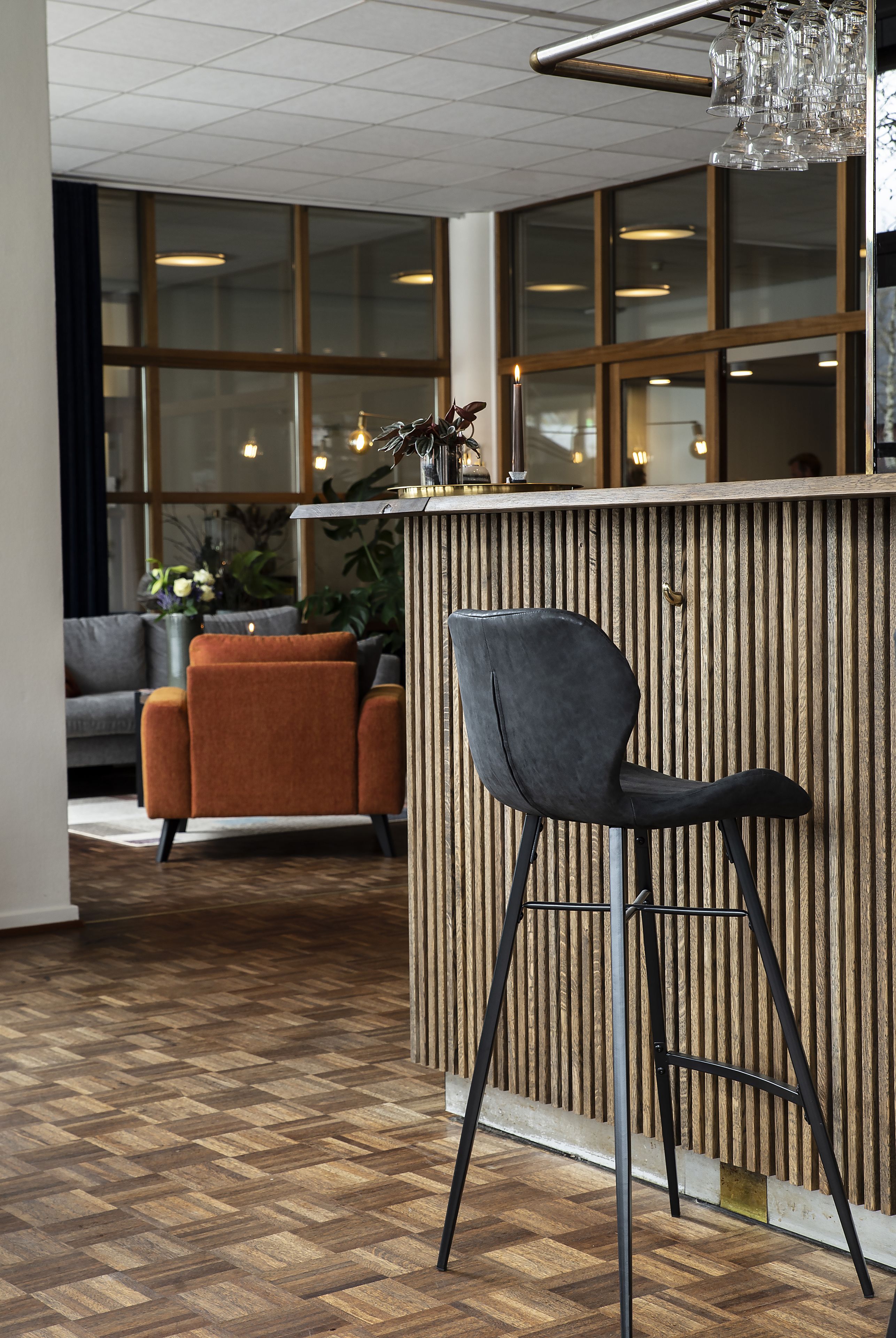 A modern interior with a wooden reception desk, a black bar stool, a grey sofa, and a patterned wooden floor.