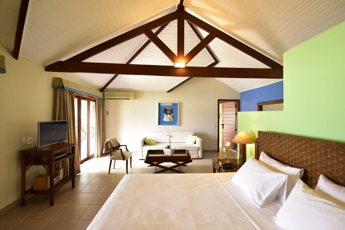 A hotel room with a large bed, a desk with a computer, a chair, a small table, a window with curtains, and a ceiling with wooden beams.