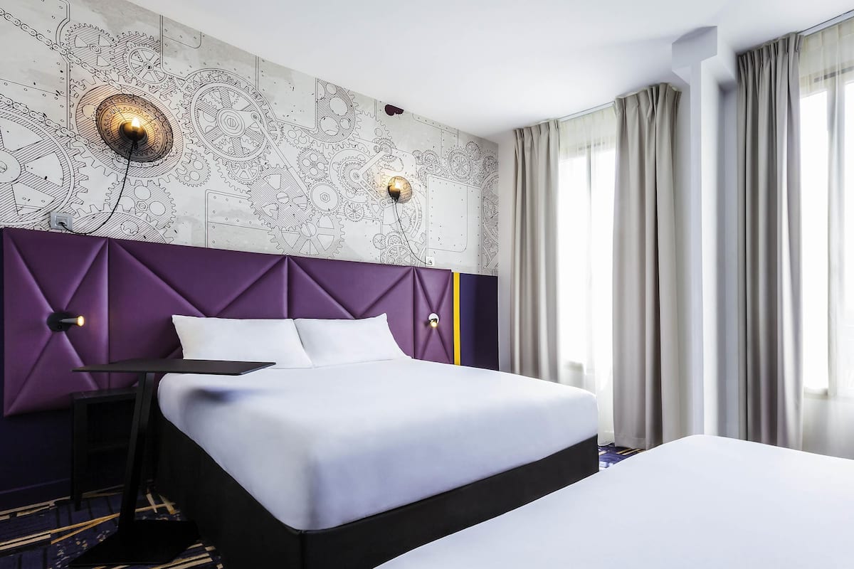 Una habitación de hotel con una cama, una mesita de noche, un muro con diseño mecánico y una ventana con cortinas.