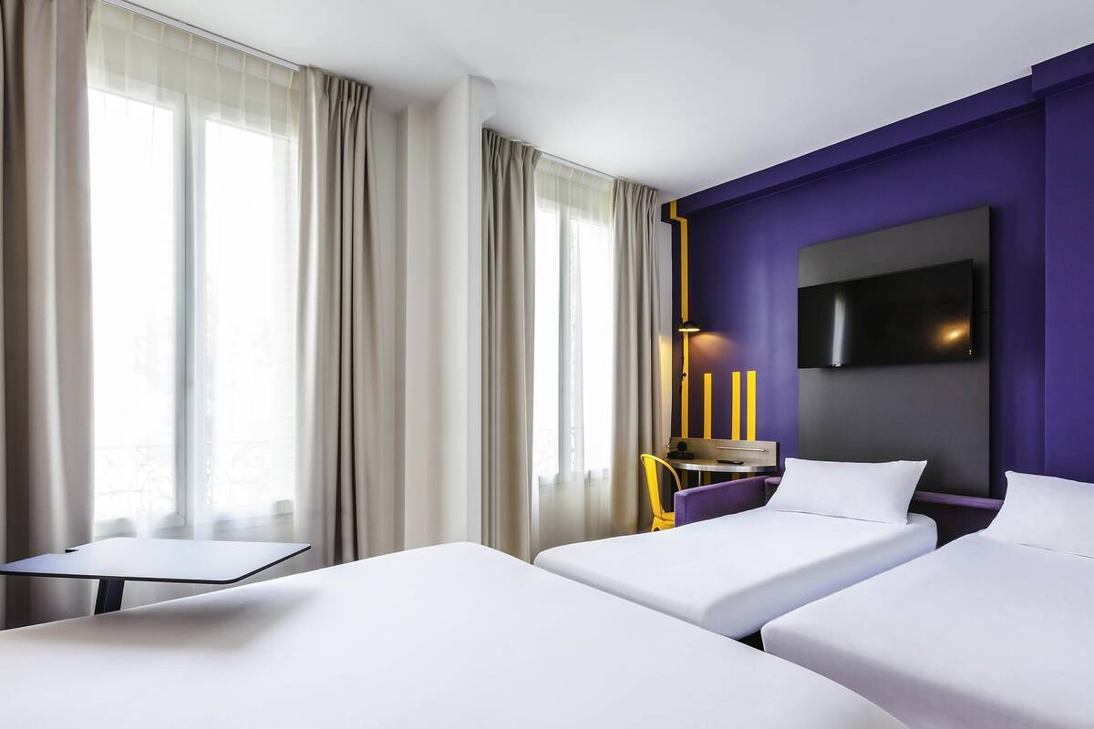 Quarto de hotel com duas camas, uma escrivaninha, uma cadeira, televisão e janelas amplas com cortinas.