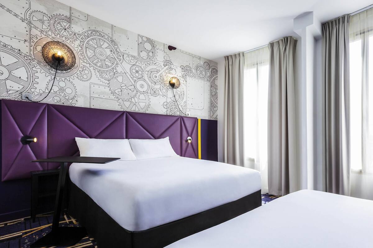 A hotel room with a large bed, a headboard with a mechanical design, two wall-mounted lights, and a window with curtains.