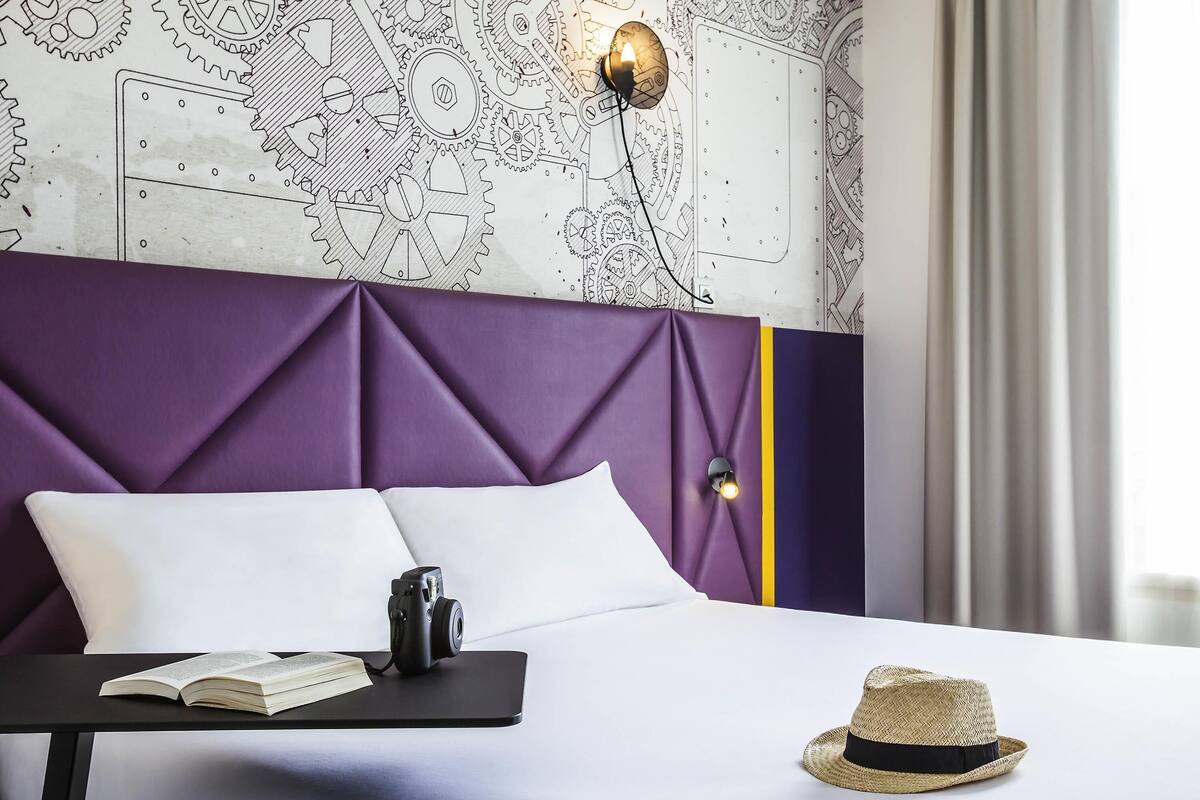 Quarto de hotel moderno com cabeceira roxa, mesa de cabeceira com uma câmera, um chapéu e um livro aberto.