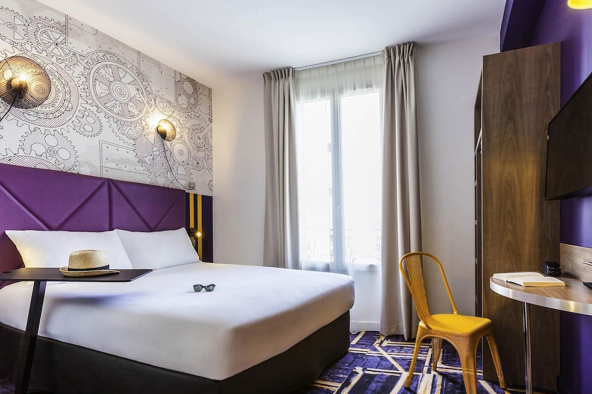 Una habitación de hotel con una cama grande, una mesita con un sombrero, una silla, una lámpara de pared y una ventana con cortinas.