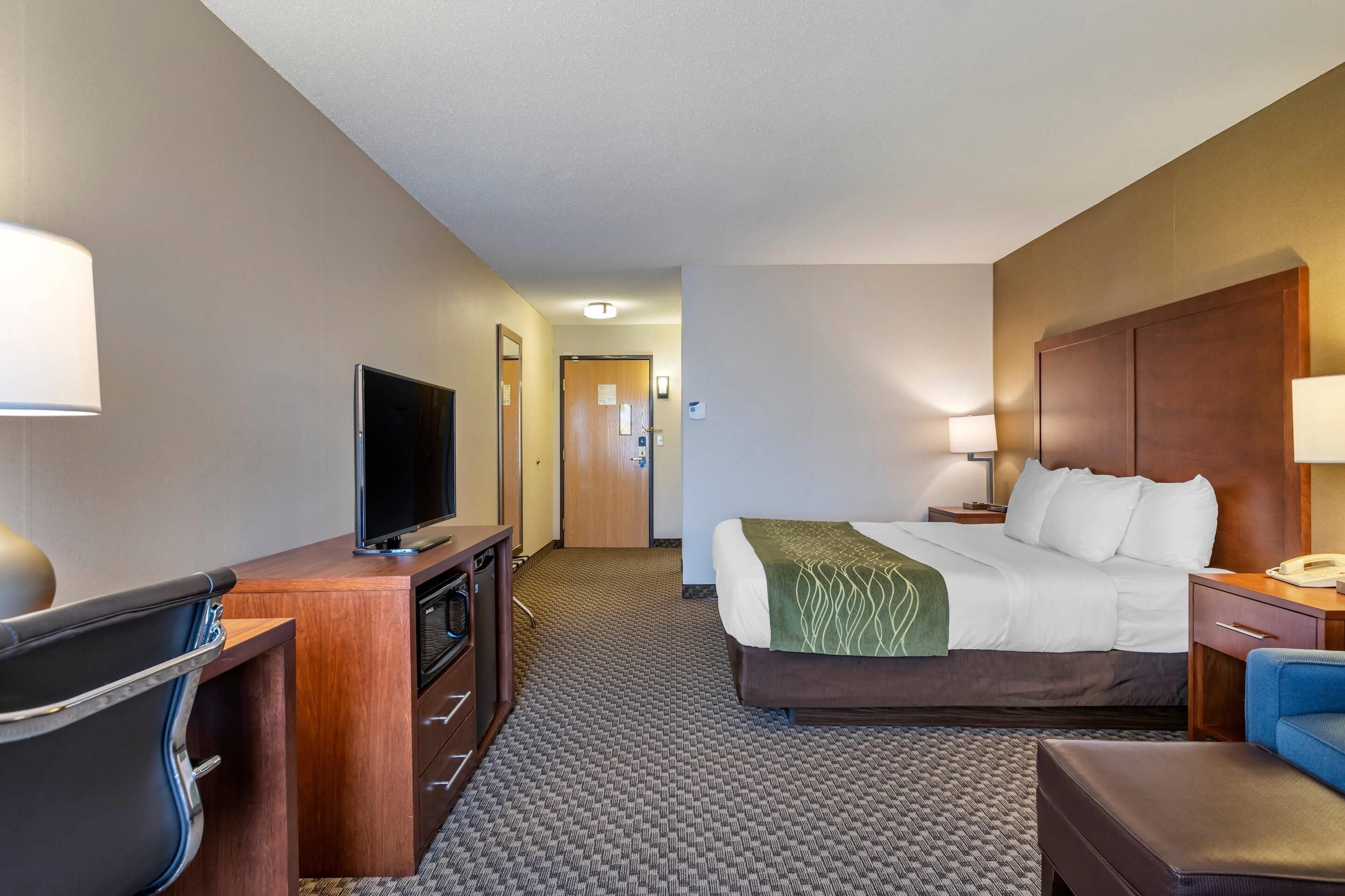 A hotel room with a bed, a desk with a television, a chair, a lamp, and a nightstand.