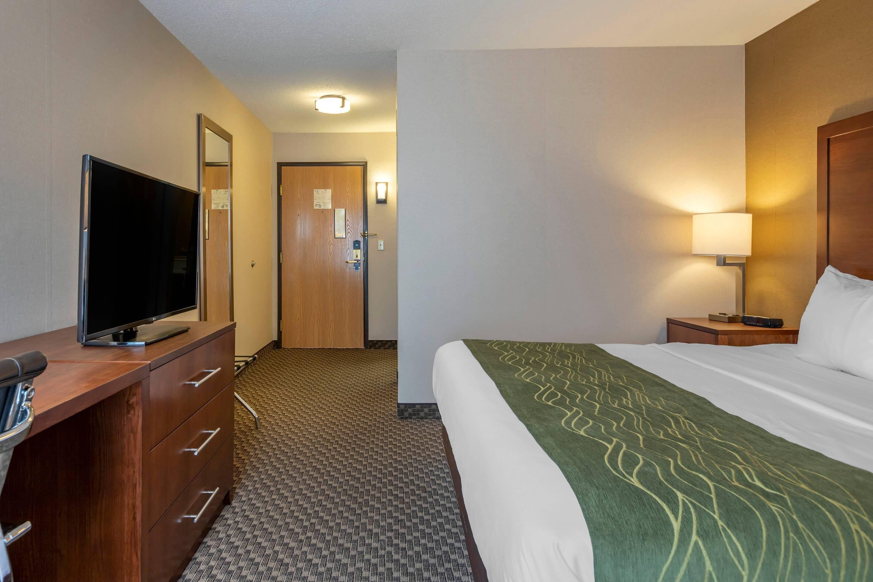 A hotel room with a bed, a TV on a wooden dresser, a nightstand with a lamp, and a door leading to another room.