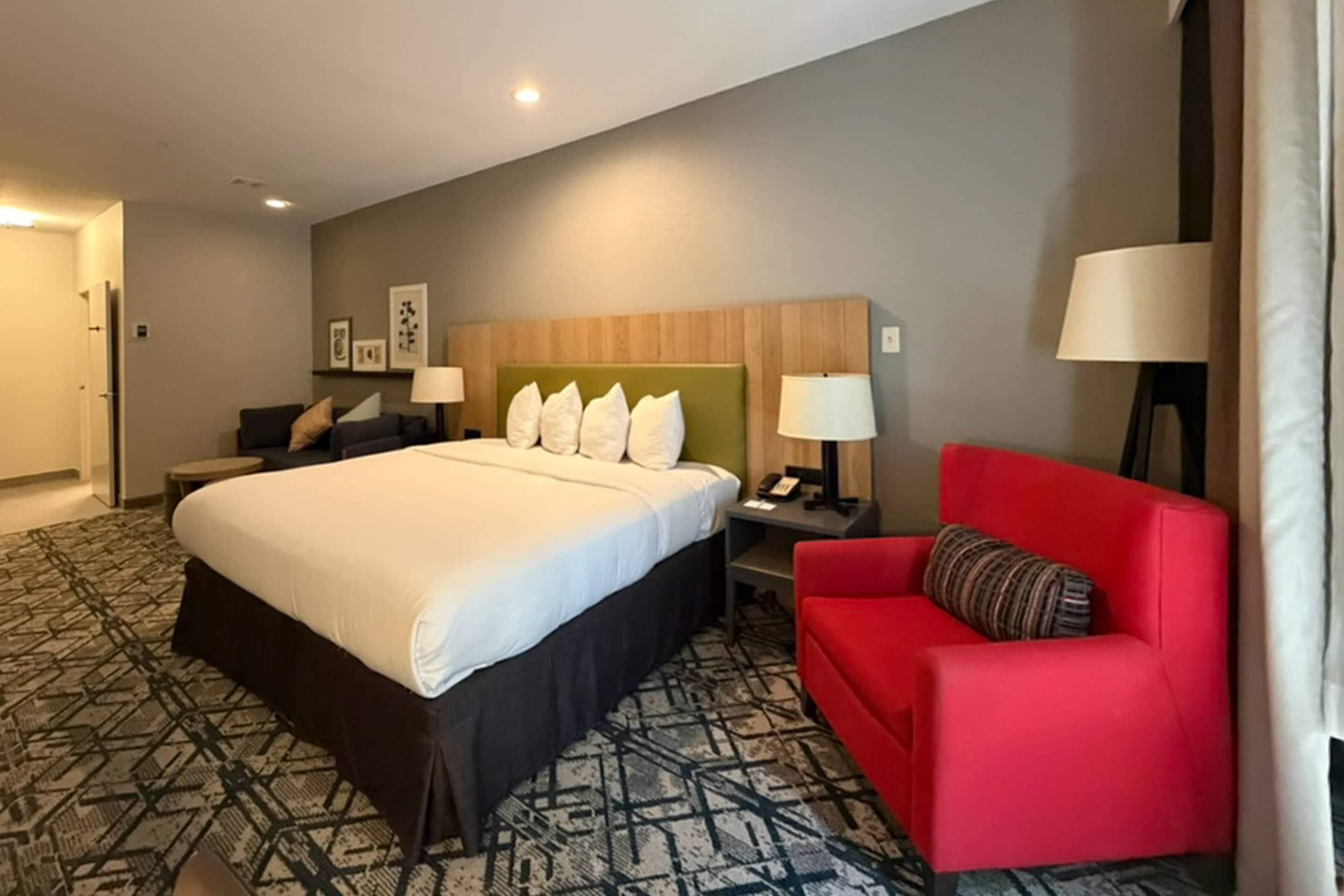 A hotel room with a large bed, a red sofa, a nightstand with a lamp, and a wall-mounted TV.