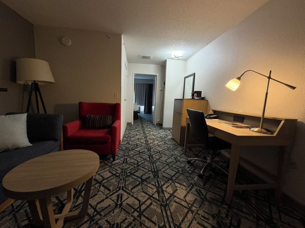A hotel room with a sofa, a red armchair, a desk with a lamp, and a patterned carpet.
